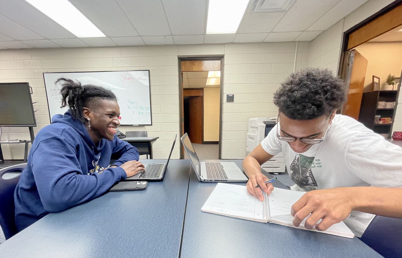 Two students laugh and study together at a table in a refurbished computer room.