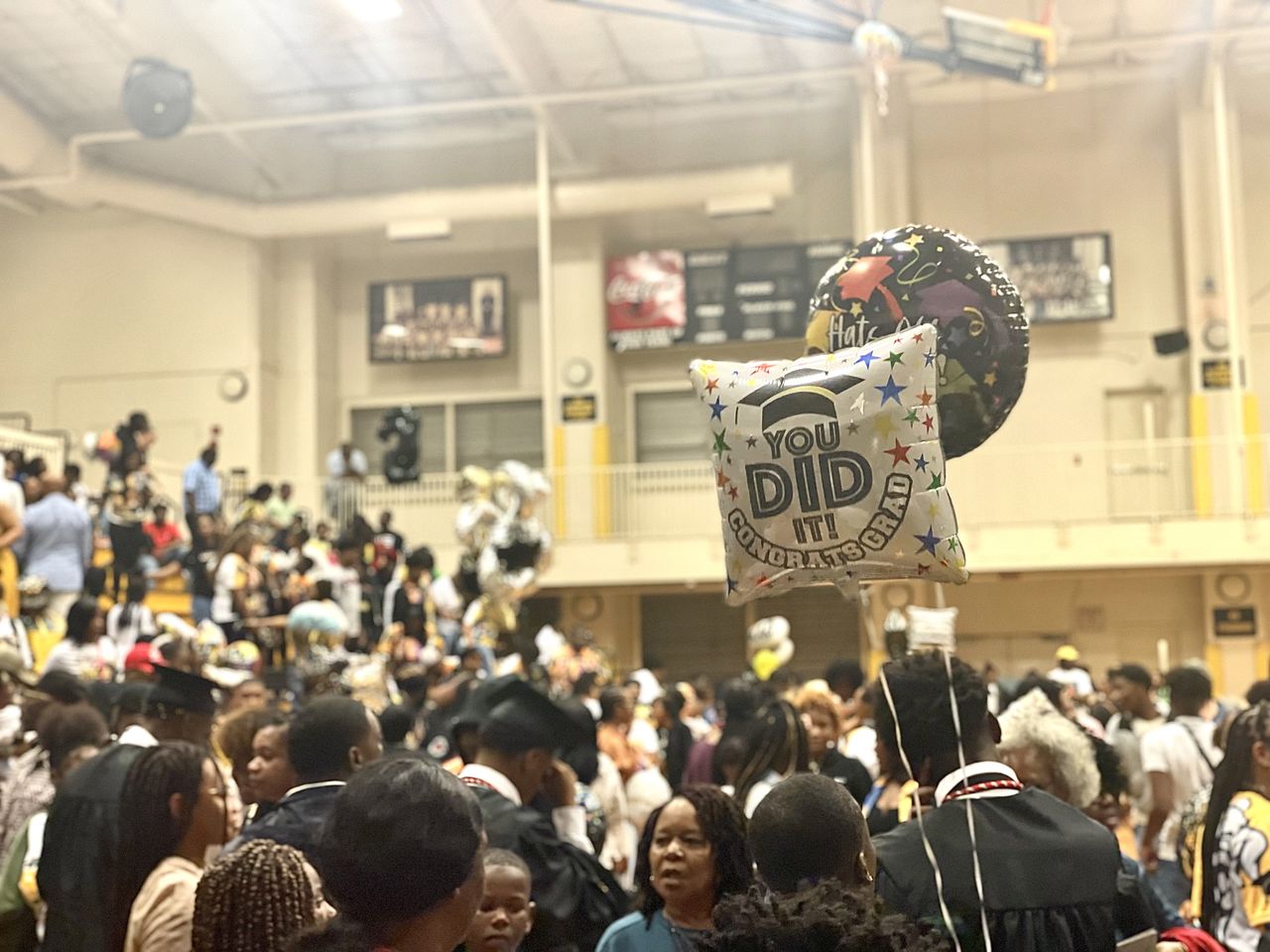 A "You Did It" balloon floats above a packed group of people in a high school gym.