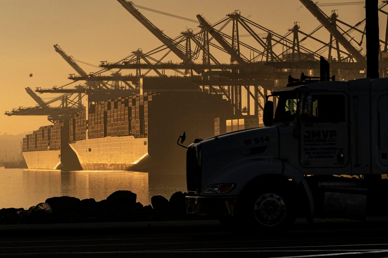 A truck arrives to pick up a shipping container in dim light as vessels are docked under cargo cranes in the background